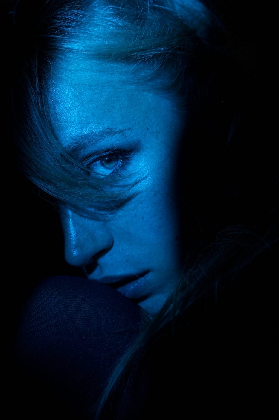 Face emerging from near darkness under cold blue light, eyes lowered, Amanda Black (@a.blck)