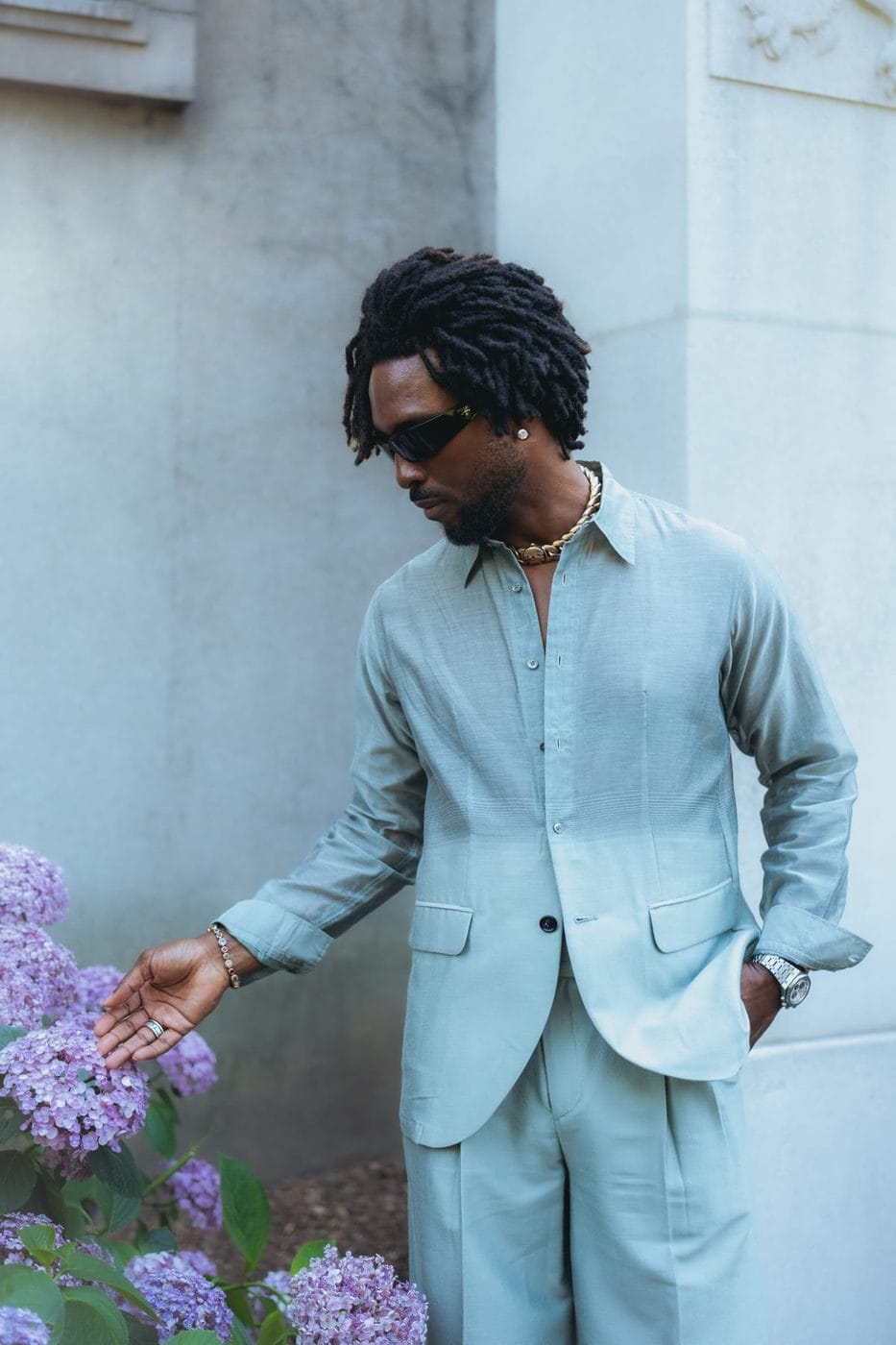 Man in a pale blue linen suit and sunglasses against a white wall, hand reaching towards hydrangeas, SAINt JHN (@saintjhn)