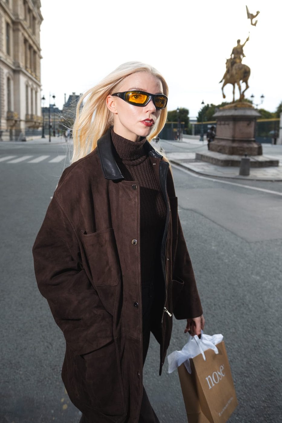 Blonde woman in an oversized brown leather coat crossing a Paris square, equestrian statue behind, Anya Taylor-Joy (@anyataylorjoy), Nose