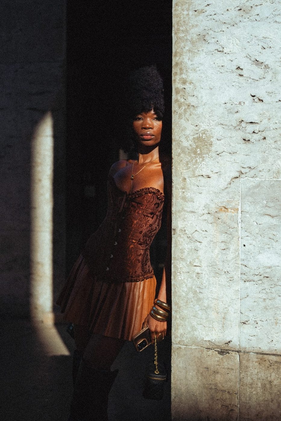 Woman in a rust corset dress standing in a shaft of light on a stone wall, Zaila (@zaila_56)