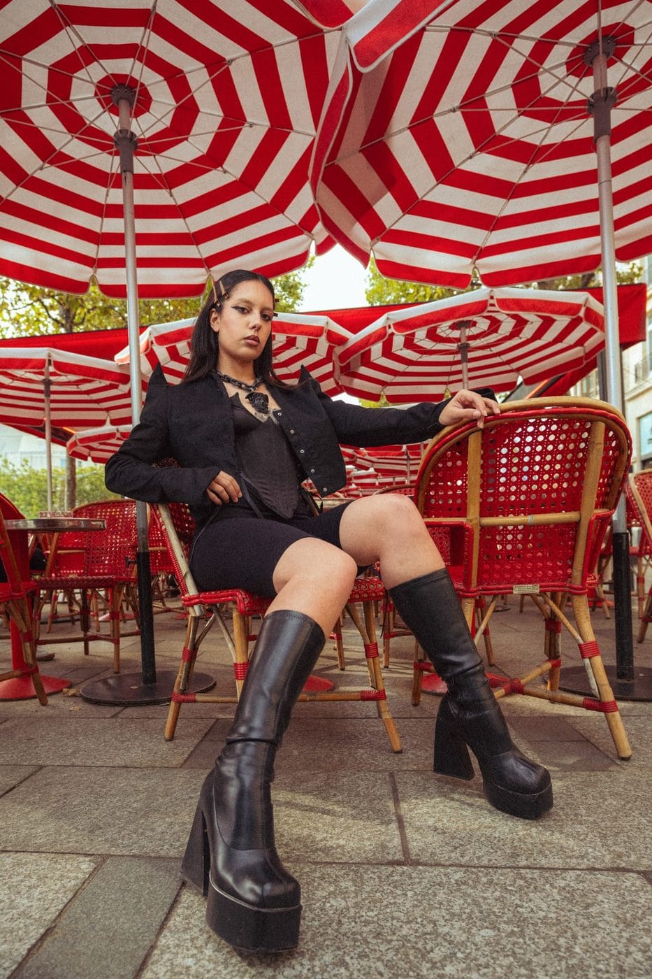 Woman in black seated under red-and-white striped café parasols, platform boots, June Viktoria Anderson (@junyeanderson)