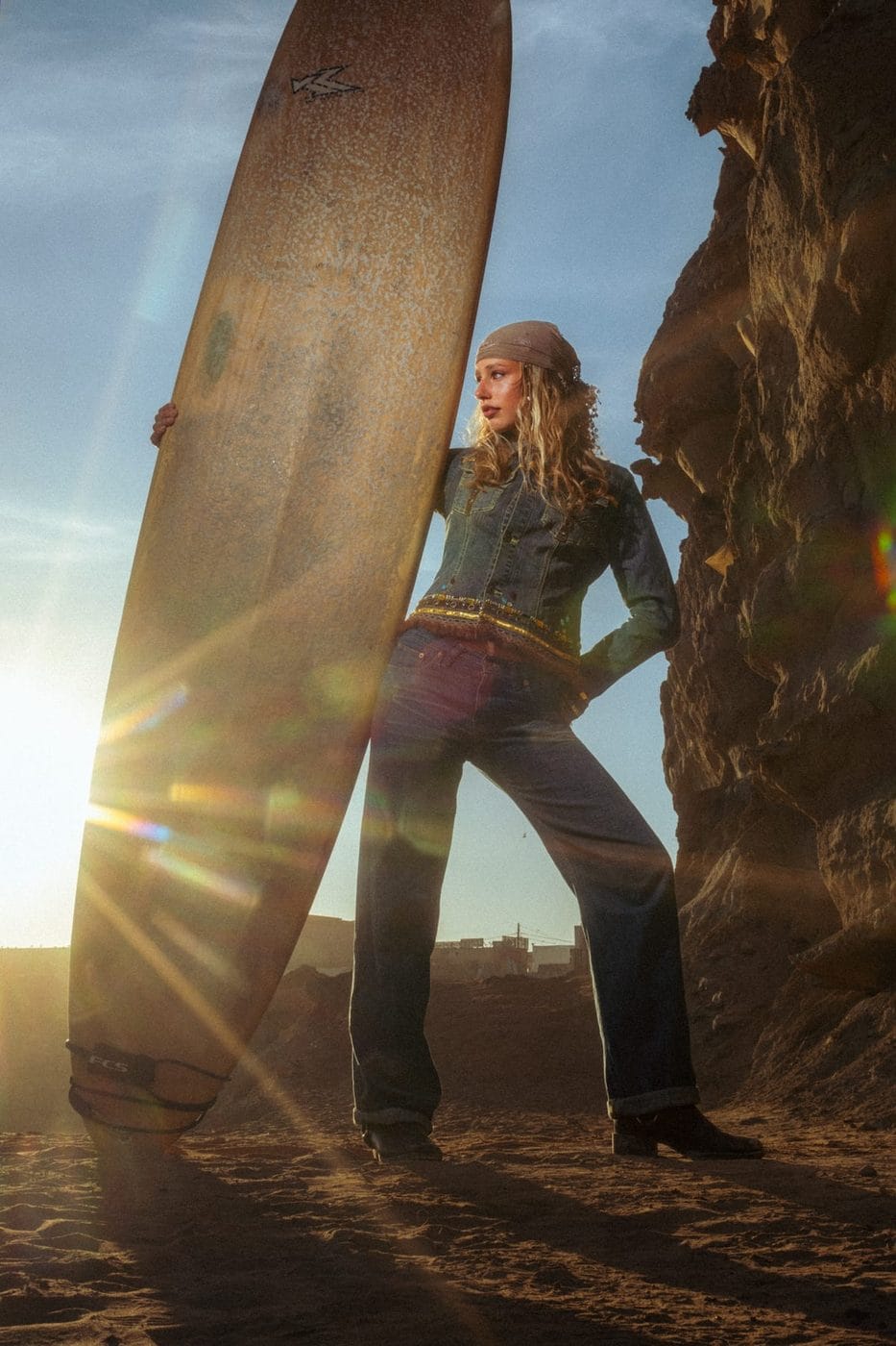 Woman in denim leaning on a surfboard at sunset, backlit by the low sun, Merle (@merlecrohn)