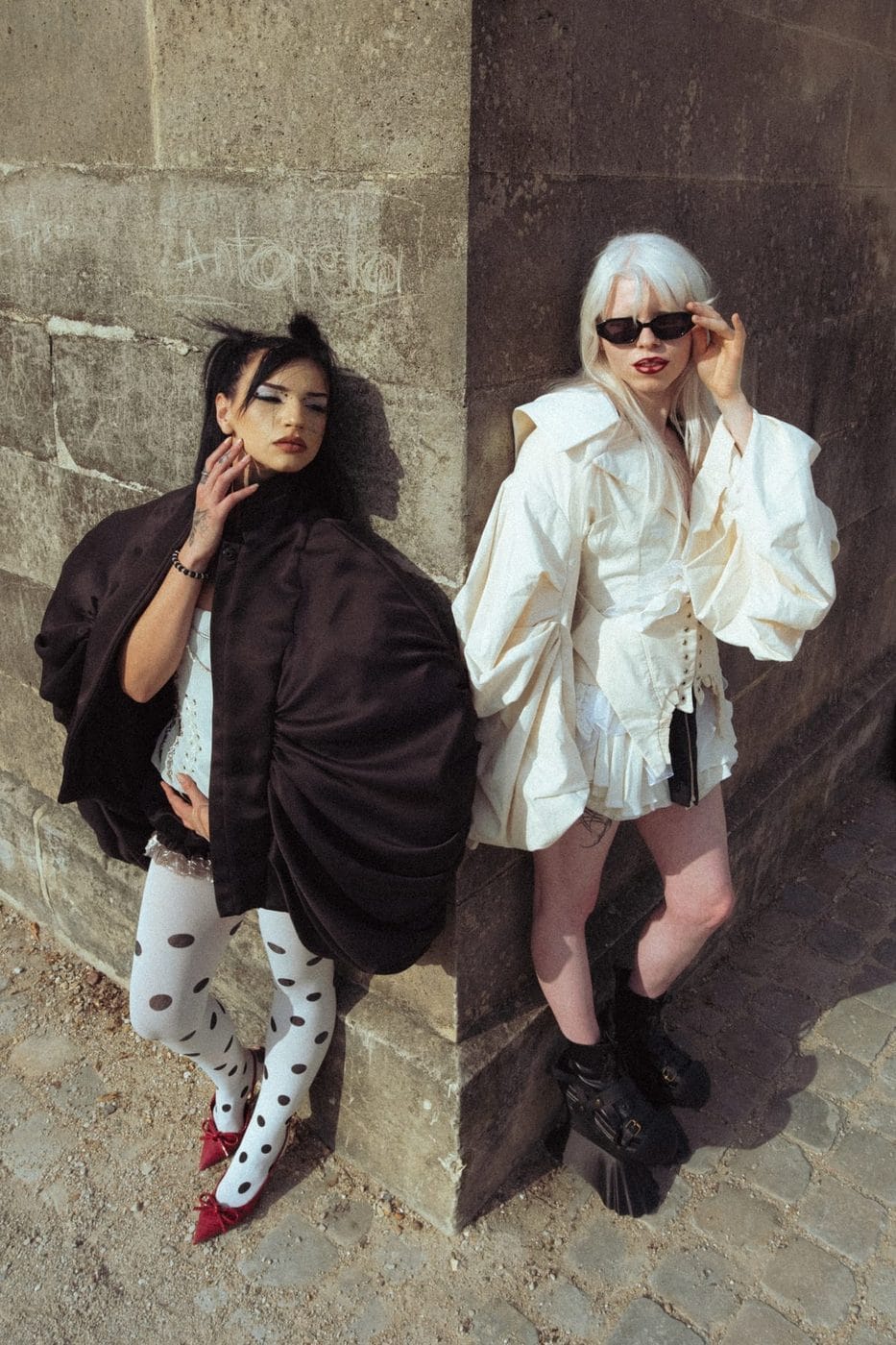 Two women against a stone wall, one in a black cape and polka-dot tights, the other in white, Gabriella Magniez (@gxbxxy) et Victoria Laurent (@2moiselle.savage), Stylisme asgoodasgold.mt, maquillage annabeth.mua