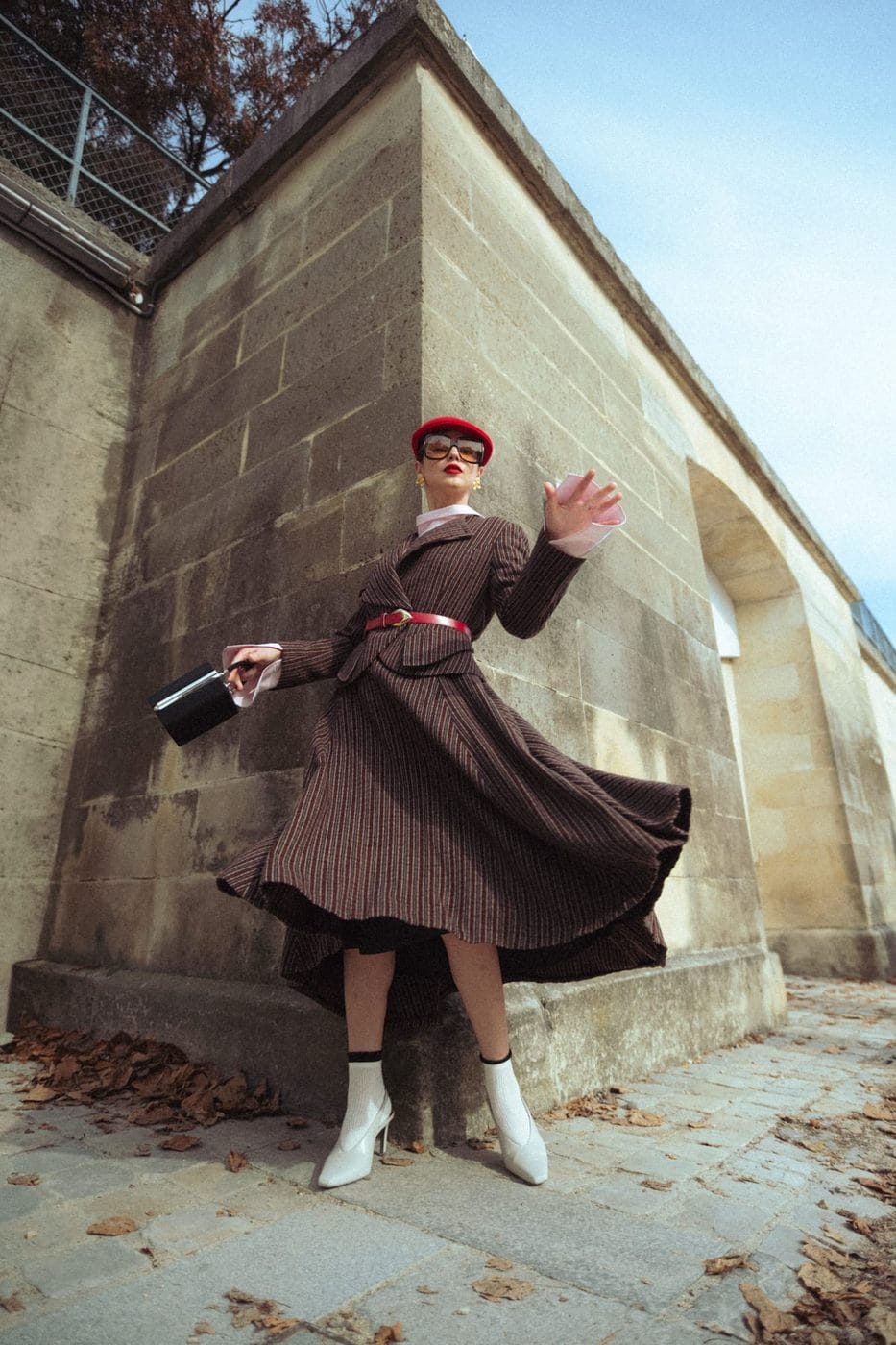 Woman in a pleated brown dress and red beret turning beside a stone wall, @olga_kuzymko