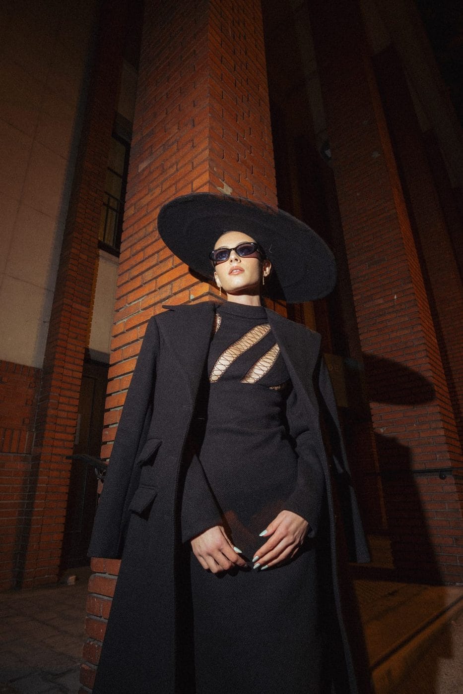 Woman in a long black coat and wide-brim hat against a brick wall at night, Amélie Diaz (@ameliediaz.co), Ses propres créations